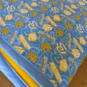 May include: A blue and yellow fabric with a repeating pattern of the words "Southern Jaguars" and a stylized jaguar head. The fabric is folded and has a yellow border.