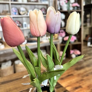 May include: A bouquet of four artificial tulips in a glass vase. The tulips are in shades of pink, peach, purple, and cream, with green stems and leaves. The arrangement is set on a wooden surface.