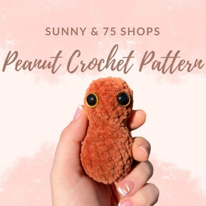 May include: A crocheted peanut plush toy with brown fur and large black eyes. The toy is being held in a hand with a light pink background. The text "Sunny & 75 Shops Peanut Crochet Pattern" is at the top of the image.