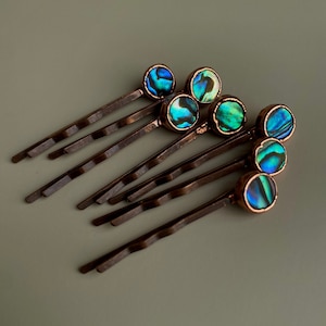 Puede incluir: Un conjunto de horquillas de color bronce, cada una coronada con una concha de abulón redonda e iridiscente. Las conchas muestran una mezcla de tonos azules, verdes y turquesas. Las horquillas están dispuestas sobre una superficie gris neutra.