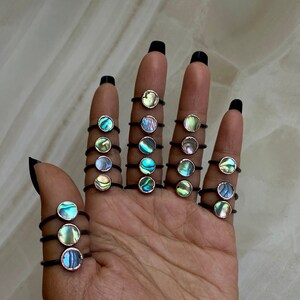 May include: A collection of rings with iridescent abalone shell insets. Each ring features a thin, black band and a circular setting. The abalone shells display a range of colors, including blues, greens, and purples, creating a unique and eye-catching design.