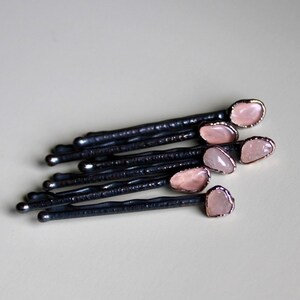 May include: A set of eight black hair pins with pink stone accents. Each pin has a small, oval-shaped pink stone set in a gold-toned metal setting.