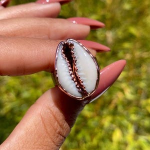 May include: A handcrafted ring featuring a white cowrie shell set in a rose gold-colored metal. The shell has a natural, textured appearance, and the ring is displayed against a blurred green background.
