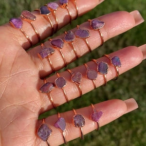 May include: A collection of 14 rose gold rings with raw purple gemstones. Each ring features a unique, irregular shaped gemstone.