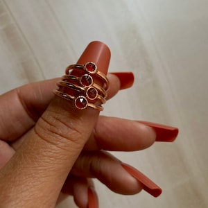 May include: Set of four gold rings with red gemstone accents. The rings are stacked on a finger.