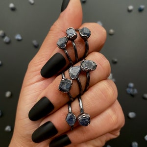May include: A set of five silver rings with raw blue sapphire gemstones. The rings are stacked on each other and are being held by a hand with black nail polish.