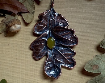 May include: A copper-coloured leaf pendant with a green gemstone centre hangs from a fine chain. The pendant is surrounded by dried oak leaves and acorns, creating a rustic, autumnal theme. The leaf design features intricate veining.