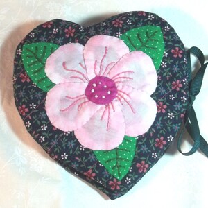以下が含まれることがあります： 大きなピンクの花のアップリケが付いた、ハート型のキルト生地アイテム。花は濃いピンク色の中心部を持ち、緑の葉に囲まれています。背景の生地は、小さな花柄のネイビーブルーです。閉じるための緑のリボンが付いています。