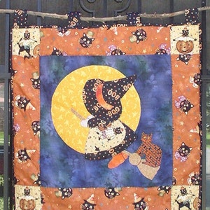 May include: A Halloween-themed quilt featuring a witch flying on a broomstick in front of a full moon. The quilt is made from orange, brown, and blue fabric and includes black cats, pumpkins, and stars. The witch is wearing a black hat and dress with the word "BOO" written on it.