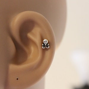 May include: A silver ear stud with a small, intricate design. The stud is worn in the cartilage of the ear.