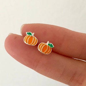 Puede incluir: Dos pequeños pendientes de calabaza naranja con contornos blancos y hojas verdes. Los pendientes se sostienen entre los dedos, mostrando su tamaño en miniatura y su tema otoñal. Un accesorio de temporada.