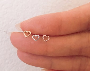 May include: Three small heart-shaped earrings in different metal finishes: gold, silver, and rose gold. The earrings are displayed on a hand.