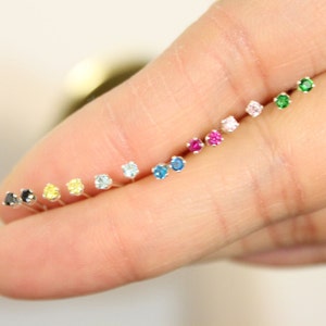 Pu&ograve; includere: Una fila di piccoli orecchini a perno d'argento con pietre preziose di diversi colori. I colori includono nero, giallo, azzurro, blu scuro, rosa, rosso e verde.