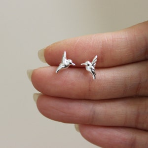 Könnte beinhalten: Ein Paar silberne Kolibri-Ohrringe. Die Ohrringe sind klein und zart und haben ein realistisches Kolibri-Design.