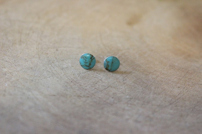 May include: A pair of turquoise stud earrings. Each earring features a round, polished turquoise stone set in a silver-colored metal backing. The earrings are displayed on a light brown wooden surface.