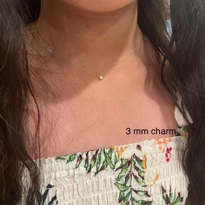 May include: A gold chain necklace with a small, clear 3 mm charm. The necklace is worn by a person with long, dark hair and a white floral top.