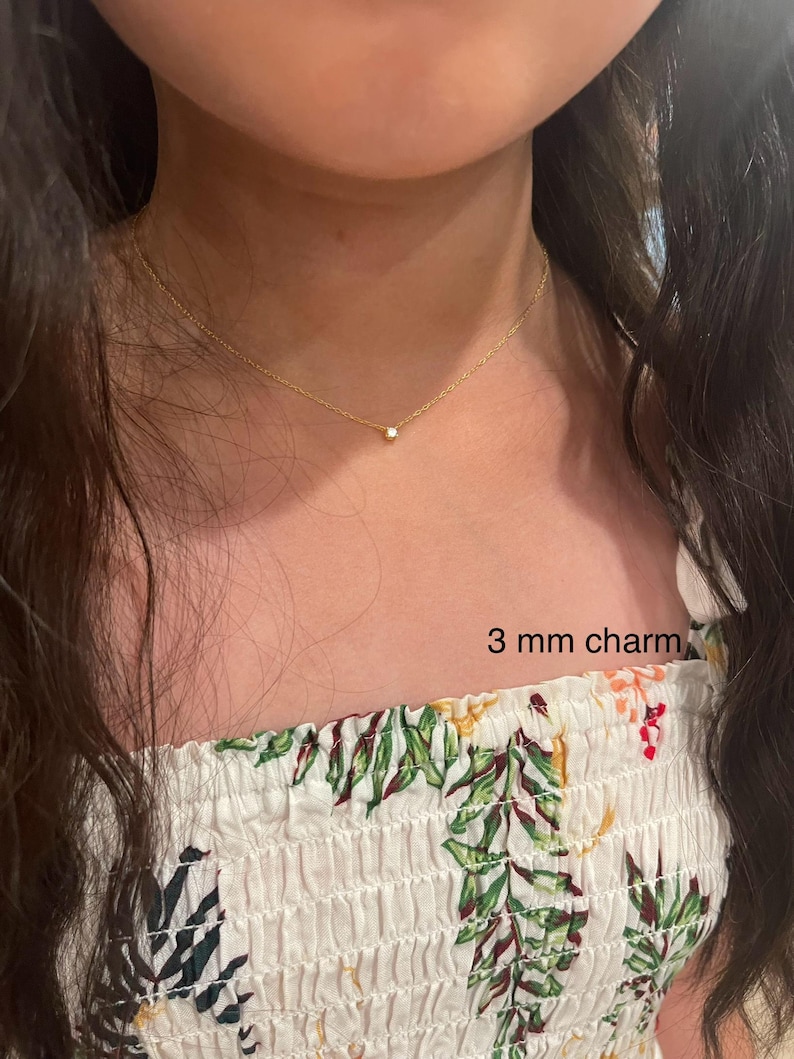 May include: A gold chain necklace with a small, clear 3 mm charm. The necklace is worn by a person with long, dark hair and a white floral top.