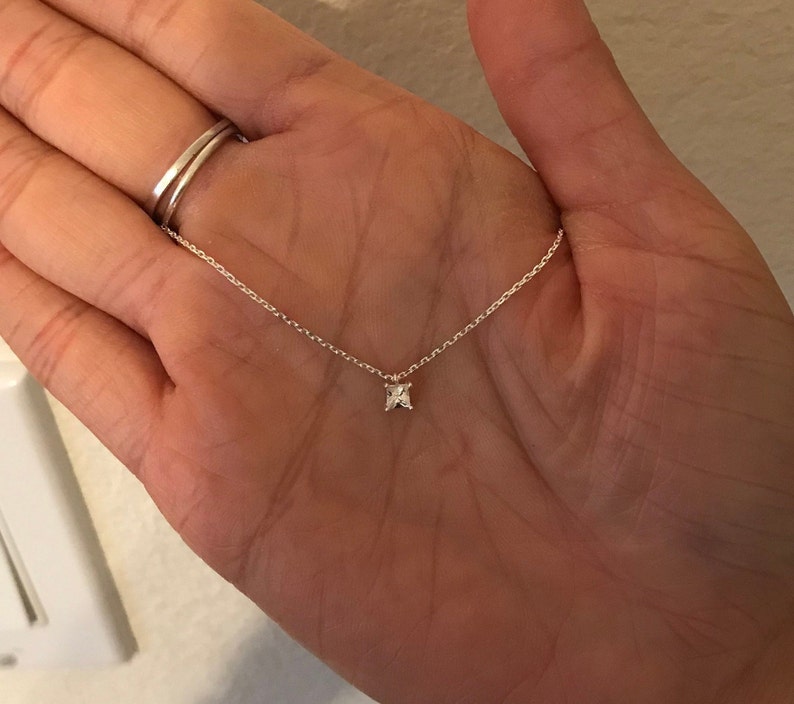 May include: Delicate silver necklace featuring a small, square-cut gemstone pendant. The pendant is centered on a fine chain, creating a minimalist and elegant design. The necklace is shown against a neutral background.