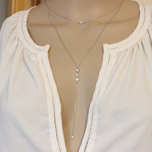 May include: A silver layered necklace with a long pendant chain featuring three small clear stones and a single stone at the bottom. The necklace is worn on a white blouse.