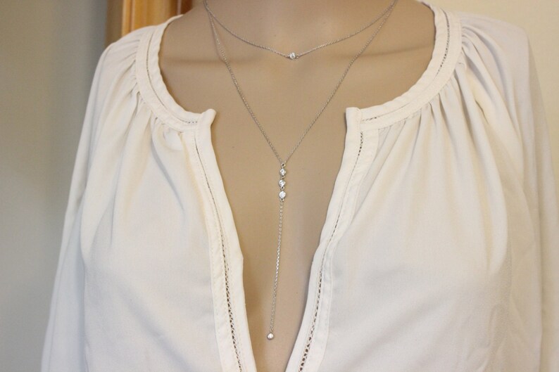 May include: A silver layered necklace with a long pendant chain featuring three small clear stones and a single stone at the bottom. The necklace is worn on a white blouse.