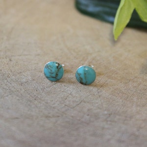 May include: A pair of turquoise stud earrings. Each earring features a round, polished turquoise stone with brown veining, set on a silver post. The earrings are displayed on a light brown wooden surface.