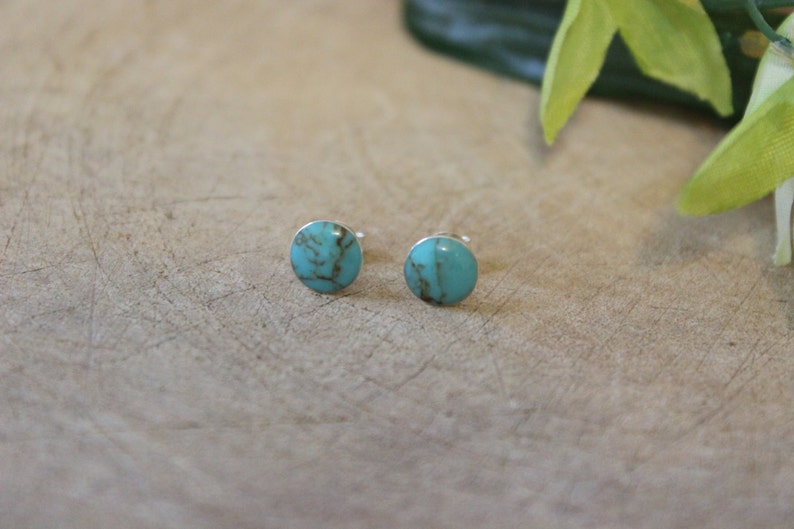 May include: A pair of turquoise stud earrings. Each earring features a round, polished turquoise stone with brown veining, set on a silver post. The earrings are displayed on a light brown wooden surface.