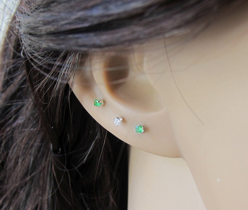 May include: Three small earrings with gemstones. The earrings are silver and have a green and clear gemstone. The earrings are worn in the earlobe.