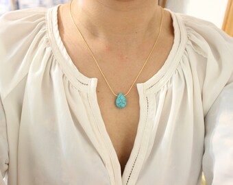 Puede incluir: Un collar de cadena dorada con un colgante de lágrima turquesa. El collar lo lleva una persona que lleva una blusa blanca.