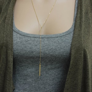 Puede incluir: Un collar largo y delgado de oro con un colgante de barra. El collar se lleva sobre un top gris sin mangas y una chaqueta de punto verde.
