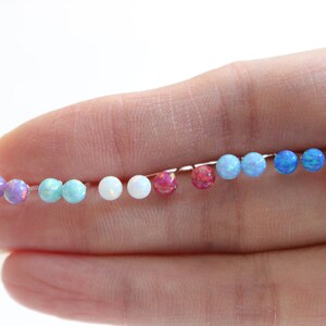 May include: A set of 10 opal stud earrings in various colors, including pink, blue, white, and turquoise. The earrings are arranged in a line on a hand.