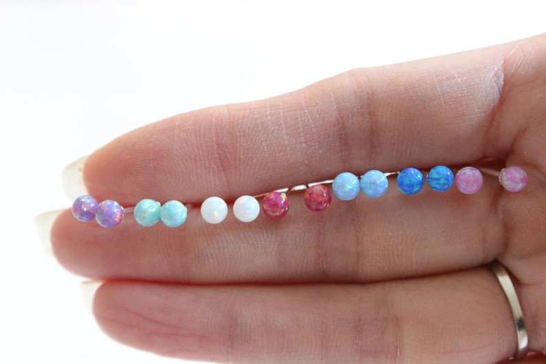 May include: A set of 10 opal stud earrings in various colors, including pink, blue, white, and turquoise. The earrings are arranged in a line on a hand.