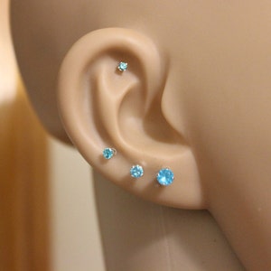 Pu&ograve; includere: Primo piano di un orecchio con quattro piccoli orecchini di pietre blu. Gli orecchini sono incastonati in argento.