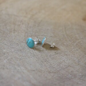 May include: A pair of turquoise stud earrings. Each earring features a polished, oval-shaped turquoise stone set in a silver backing. The earrings are displayed on a light brown, textured surface.