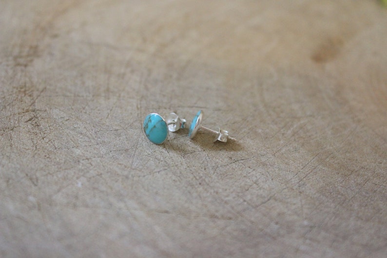 May include: A pair of turquoise stud earrings. Each earring features a polished, oval-shaped turquoise stone set in a silver backing. The earrings are displayed on a light brown, textured surface.
