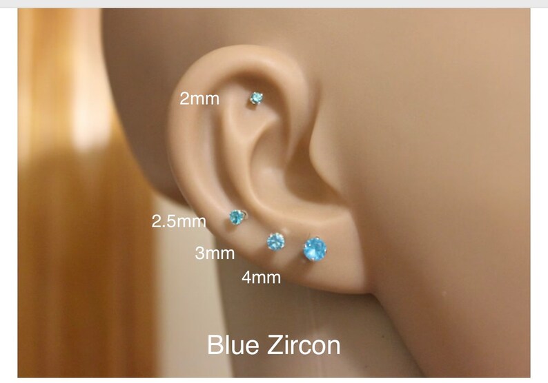 Puede incluir: Cuatro pares de pendientes de bot&oacute;n de circonita azul en diferentes tama&ntilde;os: 2 mm, 2,5 mm, 3 mm y 4 mm. Los pendientes se muestran en la oreja de un maniqu&iacute;.