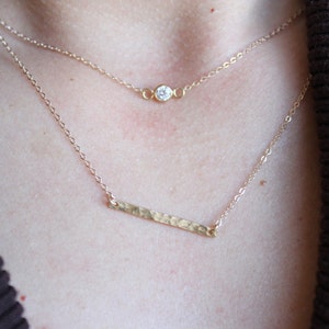 Op de afbeelding: Een gouden ketting met een klein, rond, helder edelsteen hanger en een langere, gehamerde gouden staaf hanger.