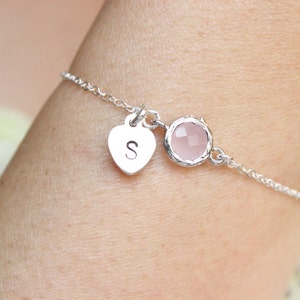 May include: A delicate silver bracelet featuring a heart-shaped charm with the letter "S" and a round, light pink gemstone. The bracelet is on a chain link design.