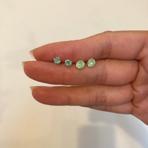 May include: Four pairs of small, round, green opal stud earrings. The earrings are arranged in a row on a finger.