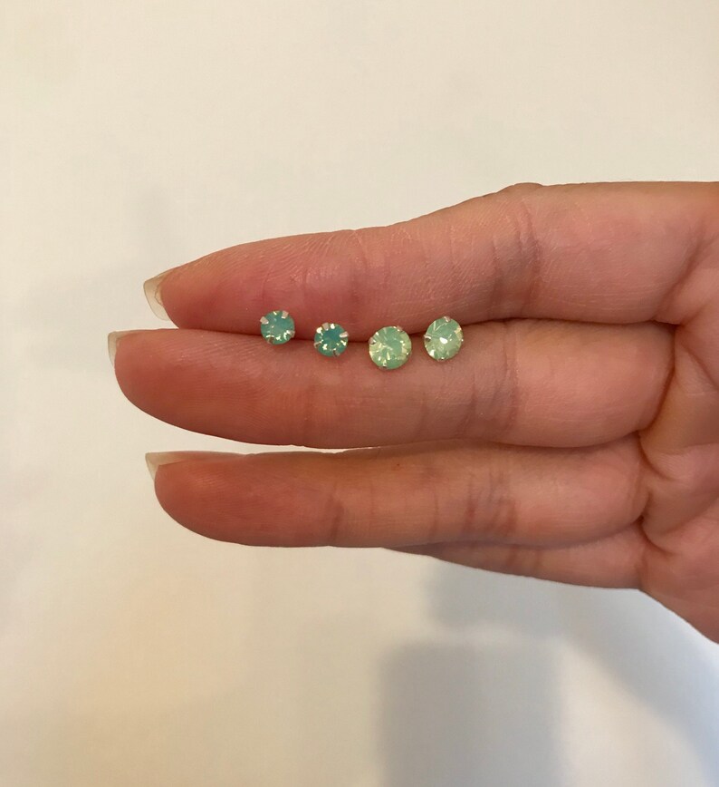 May include: Four pairs of small, round, green opal stud earrings. The earrings are arranged in a row on a finger.