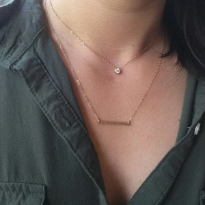 Op de afbeelding: Een gouden ketting met een klein rond hangertje en een dunne staafhanger.
