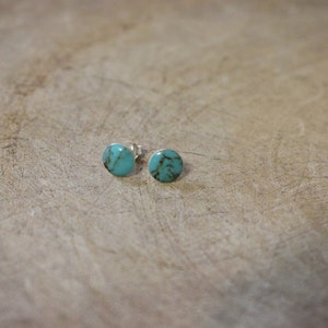 May include: A pair of turquoise stud earrings. Each earring features a round, polished turquoise stone set in a silver backing. The earrings are displayed on a light brown, textured surface.