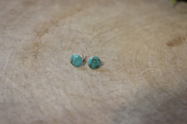 May include: A pair of turquoise stud earrings. Each earring features a round, polished turquoise stone set in a silver backing. The earrings are displayed on a light brown, textured surface.