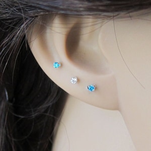 May include: Three small stud earrings with clear and blue gemstones. The earrings are worn in the earlobe and helix piercing.