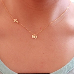 Puede incluir: Un collar de cadena dorada con un pequeño dije de cruz y dos dijes de iniciales. Las iniciales son "M" y "H".