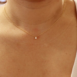 May include: A delicate gold necklace featuring a small, square, clear gemstone pendant. The necklace is displayed on a person's chest, showcasing the minimalist design and subtle elegance of the jewelry.