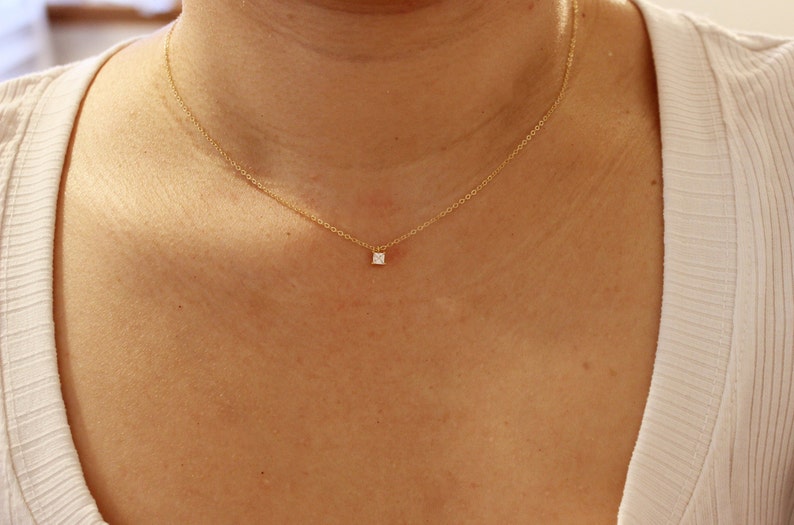May include: A delicate gold necklace featuring a small, square, clear gemstone pendant. The necklace is displayed on a person's chest, showcasing the minimalist design and subtle elegance of the jewelry.