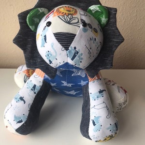 May include: A patchwork lion stuffed animal with a blue, white, and green striped mane. The lion has black button eyes and is made from a variety of fabrics with different patterns.