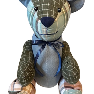 May include: A handcrafted teddy bear with a patchwork design. The bear has a blue and green plaid pattern on its head, arms, and feet, with a solid blue body. It has a dark blue bow and black button eyes. The bear is sitting upright.