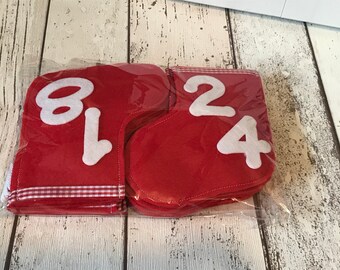 May include: A stack of red felt Christmas stockings with white numbers and accents. The numbers 18 and 24 are visible. Each stocking has a white and red checkered trim. The stockings are packaged in a clear plastic bag.