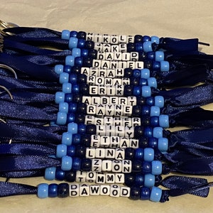 May include: A set of 18 blue and white beaded keychains with names spelled out on each keychain. The keychains are made with blue and white beads and have a blue ribbon attached to each keychain.
